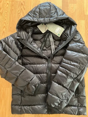 Svart dunjacka från Canada Goose - Snygg svart dunjacka från Canada Goose med huva och dragkedja framtill. Jackan har två sidofickor med dragkedja, quiltad design och logotypmärke på ärmen. Perfekt för kalla vinterdagar och riktigt varm tack vare dunfyllningen.