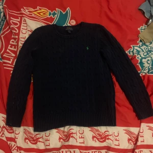 Mörkblå kabelstickad tröja Polo - Snygg mörkblå kabelstickad tröja från Polo Ralph Lauren med rund halsringning och den klassiska gröna logon broderad på bröstet. Tröjan har långa ärmar och ribbade muddar vid ärmslut och nederkant. Perfekt för en stilren och avslappnad look.