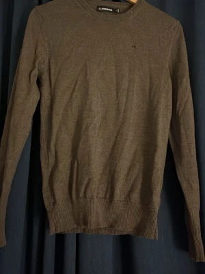 Brun tröja från J.Lindeberg - Mjuk och tunnstickad brun tröja från J.Lindeberg med rund halsringning. Tröjan har långa ärmar och ribbade muddar vid ärmslut och nederkant. Perfekt för lager-på-lager och enkel att matcha med jeans eller chinos.