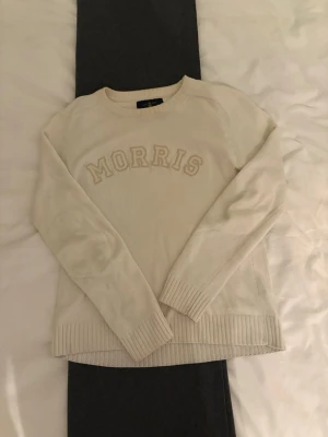 Vit stickad tröja från Morris - Snygg vit stickad tröja från Morris med broderad logga framtill. Tröjan har rund halsringning, långa ärmar och ribbade muddar vid ärmslut och nederkant. Perfekt för en stilren och avslappnad look.