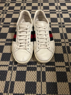 Gucci vita sneakers med röd-blå rand - Snygga vita sneakers från Gucci i läder med klassiska röd-blå ränder på sidan och svart detalj vid hälen. Skorna har vita snören och låg profil som ger en clean look. Perfekta för dig som vill ha en stilren och lyxig sneaker med ikonisk design.