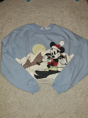 Ljusblå Mickey Mouse tröja H&M - Säljer en ljusblå croppad sweatshirt från H&M med stort tryck av Mickey Mouse som åker skidor framför berg och sol. Tröjan har långa ärmar, ribbade muddar och rund hals. Perfekt för dig som gillar Disney och vill ha en chill vinterlook.
