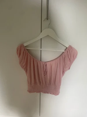 Ljusrosa offshoulder topp - Säljer en ljusrosa topp med offshoulder och korta puffärmar. Toppen har en söt knytning framtill och smockad nederkant som ger en snygg passform. Materialet känns mjukt och lätt.