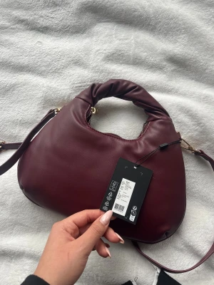 Vinröd axelväska i skinnimitation - Helt oanvänd, med prislapp. Snygg burgundy axelväska med rundad form och mjukt, puffigt handtag. Väskan har guldfärgade detaljer och är från Pieces . Tillverkad i skinnimitation och har ett innerfack med dragkedja. Perfekt storlek för det viktigaste 