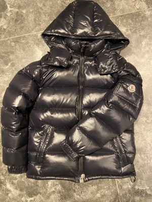 Svart Moncler pufferjacka med huva - Pris kan diskuteras. 8,8/10 skick, fråga om fler bilder om du är intresserad. Svart pufferjacka från Moncler med glansig finish och huva. Jackan har dragkedja framtill, två sidofickor med dragkedja och Moncler-logga på ärmen. Insidan har ett coolt serietryck och en innerficka med dragkedja. Perfekt för kalla dagar.