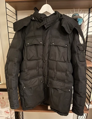Svart pufferjacka från Belstaff - Svart pufferjacka från Belstaff med coolt emblem på ärmen och flera stora fickor med knappar. Jackan har hög krage, avtagbar huva och quiltad design som ger extra värme. Perfekt för kalla dagar och har en snygg, urban look. Jackan kan ev behöva en tvätt men det får ev köpare avgöra. Priset är justerat för detta. Inköpt på NK i Göteborg men väldigt sparsamt använd. 