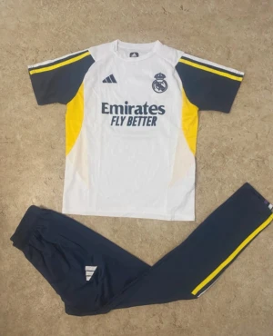 Real Madrid fotbollströja Adidas - Säljer en Real Madrid fotbollströja från Adidas i vitt med mörkblå och gula detaljer. Tröjan har korta ärmar och klubbens logga samt Emirates-tryck på bröstet. Materialet är lätt och andas, perfekt för träning eller match.
