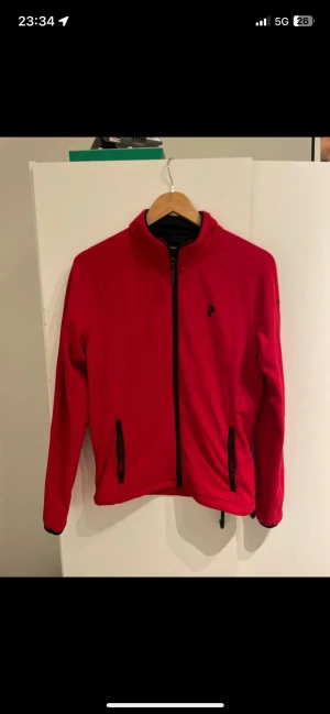 Röd vindjacka med dragkedja - Snygg röd vindjacka från Peak Performance med svart dragkedja och två fickor med dragkedja. Jackan har lång ärm och sportig look, perfekt för dig som gillar stilren design och funktion. Liten logga på bröstet.