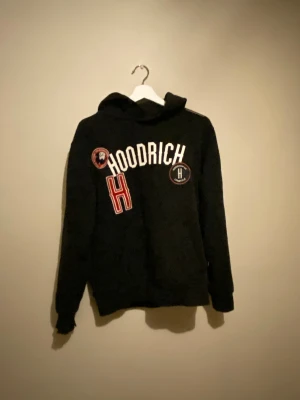 Hoodrich - Svart hoodie från Hoodrich med broderade detaljer och tryck i vitt och rött framtill. Tröjan har huva och långa ärmar, perfekt för en avslappnad streetwear-look.