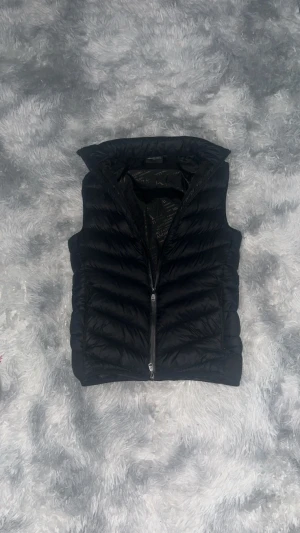 Svart dunväst Alessandro Zavetti XS - Svart dunväst från Alessandro Zavetti i storlek XS. Västen har quiltad design med horisontella sömmar, hög krage och dragkedja framtill. Insidan är fodrad med mönstrat tyg med logotyptryck. Perfekt för lager-på-lager och streetwear.