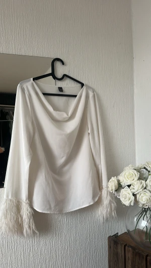Vit blus med fjädrar - Säljer en vit blus med långärmade ärmar och snygg draperad halsringning. Ärmsluten har fluffiga vita fjädrar som ger en extra cool touch. Perfekt för dig som gillar att sticka ut med detaljer. Blusen är i ett mjukt, slätt material.
