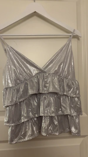 Glittrig silver volang-linnetopp - Säljer en glittrig silverfärgad linnetopp med flera lager volanger och smala axelband. Toppen har en snygg omlott-v-ringning och är croppad i modellen. Aldrig använd pågrund av att jag har lite större bröst o fick inte på mig tröjan men perfekt om du har kanske A-C kupa