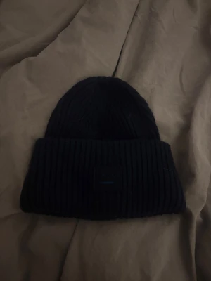 Mörkblå ribbad mössa från Acne Studios❤️ - Säljer en mörkblå ribbstickad mössa från Acne Studios med den klassiska face-loggan framtill. Mössan är tjock och har uppvikt kant, perfekt för kalla dagar. Materialet är mjukt och värmande ull, riktigt snygg och enkel att matcha med allt.