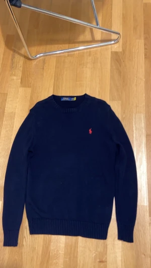 Marinblå stickad tröja Polo Ralph Lauren - Snygg marinblå stickad tröja från Polo Ralph Lauren med klassisk röd logga broderad på bröstet. Rund hals och ribbade muddar vid ärmslut och nederkant. Perfekt för dig som gillar stilrena och tidlösa plagg. storlek S men passar även M, pris kan diskuteras 