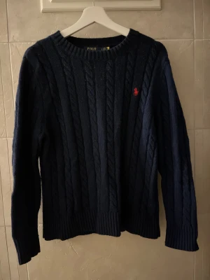 Mörkblå kabelstickad tröja Polo Ralph Lauren - Snygg mörkblå kabelstickad tröja från Polo Ralph Lauren med klassisk röd broderad logga på bröstet. Tröjan har rund hals, ribbade muddar och lång ärm. Perfekt för dig som gillar stilrena och tidlösa plagg.