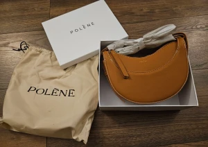 Brun axelväska från Polène - Snygg axelväska från Polène i en varm brun färg med halvmåneformad design. Väskan är tillverkad i strukturerat skinn och har vita kontrastsömmar samt en dragkedja upptill. Perfekt storlek för det viktigaste och en stilren look.