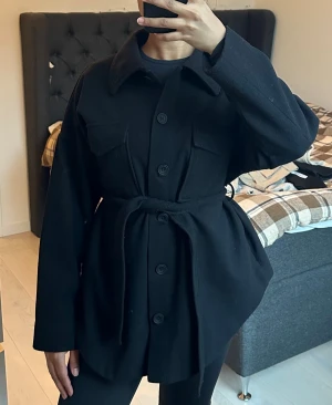 Svart overshirt med bälte och knappar - Snygg svart overshirt med kragar, knappar framtill och två bröstfickor. Jackan har ett matchande bälte i midjan för en mer markerad siluett. Långärmad och i ett mjukt ull-liknande material som passar perfekt till höst.