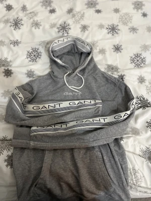 Grå hoodie från GANT med logoband - Säljer en grå hoodie från GANT med vit logoband längs ärmarna och huvan. Tröjan har huva med snörning och ribbade muddar. GANT-logga broderad på bröstet. Perfekt för chill dagar och streetstyle.