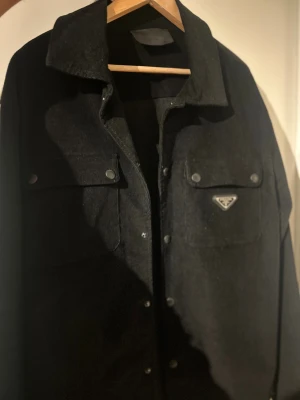 Svart manchester overshirt från Prada - Svart overshirt från Prada i mjuk manchester med klassisk krage och två stora bröstfickor med tryckknappar. Snyggt metallmärke på ena fickan och diskreta detaljer. Perfekt för dig som gillar stilrena och exklusiva plagg. Bor i Stockholm men postar den också! 