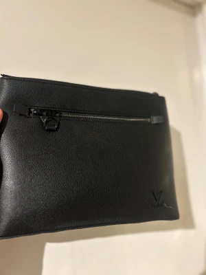 Svart kuvertväska Louis Vuitton skinn - Stilren svart kuvertväska från Louis Vuitton i exklusivt skinn. Väskan har en diskret LV-logga i hörnet och en praktisk dragkedja framtill. Perfekt för dig som vill ha en clean och lyxig accessoar med plats för det viktigaste.