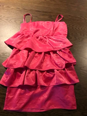 Rosa volangklänning med avtagbara axelband - Säljer en kort rosa klänning med tunna avtagbara axelband och flera lager volanger framtill. Materialet har en lätt glansig finish.            Klänningen är i storlek 36, men jag skulle säga att den är liten i storleken.