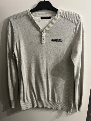 Grå ribbad tröja från LCW Casual - Snygg ljusgrå tröja från LCW Casual med ribbade detaljer på axlar, krage och muddar. V-ringad hals med tre knappar och diskret tryck på bröstet. Långärmad och tillverkad i mjuk bomullsmix som är skön mot huden.