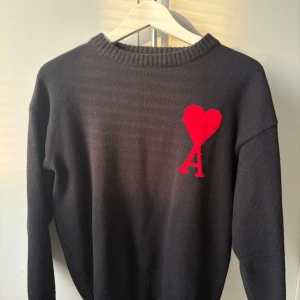 Svart stickad tröja från Ami Paris - Svart stickad tröja från Ami Paris med röd hjärtlogga och bokstaven A på bröstet. Klassisk rund halsringning och långa ärmar. Perfekt för dig som gillar stilrena plagg med en twist. Materialet är mjukt och skönt.
