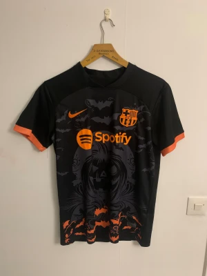 FC Barcelona Halloween fotbollströja - Säljer en FC Barcelona x Nike fotbollströja med Halloween-tema. Svart bas med orange detaljer och tryck, bland annat fladdermöss, pumpa och klubbmärke. Kortärmad modell i lätt, ventilerande material. Spotify-logga på bröstet och engineered-märke nedtill.