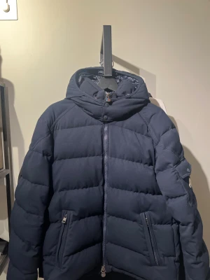 Mörkblå pufferjacka från Moncler - Säljer en Montgenevre Flannel Down Jacket Navy från Moncler med huva och snygga detaljer som dragkedja och fickor med zip. Den är i storlek 3 och passar M/L Jackan har ett quiltat mönster och är fodrad med glansigt material på insidan. Perfekt för kalla dagar och har en clean, modern look.