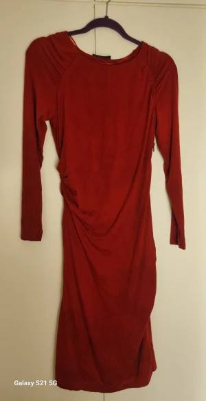 Röd långärmad klänning med dragkedja - Snygg röd långklänning med lång ärm och cool rynkning på sidan. Klänningen har en synlig dragkedja längs sidan som ger en edgy vibe. Materialet känns mjukt och har en lätt glans. Passformen är normal och klänningen har en stilren, figurnära siluett.