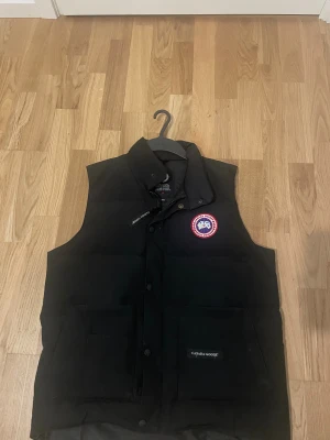 Svart dunväst från Canada Goose M - Svart dunväst från Canada Goose med klassisk logotyp på bröstet. Västen har hög krage, två stora fickor framtill och knäppning med både dragkedja och tryckknappar. Perfekt för kalla dagar och har en clean, stilren look.
