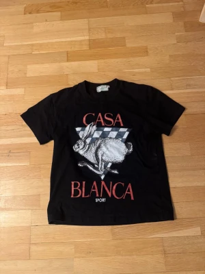 Svart Casablanca t-shirt med tryck - Cool svart t-shirt från Casablanca Tennis Club i bomull. Framsidan har ett stort tryck med en springande hare, schackrutigt mönster och röd text 'CASA BLANCA SPORT'. Klassisk passform och korta ärmar, perfekt för dig som gillar statement-plagg.