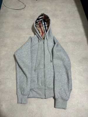 Grå Burbbery kofta med kedja  - Snygg grå hoodie från Burberry med klassiskt rutigt foder i huvan. Tröjan har dragkedja framtill, snörning vid huvan och ribbade muddar. Perfekt för dig som gillar stilrena detaljer och vill ha en hoodie med exklusiv känsla.