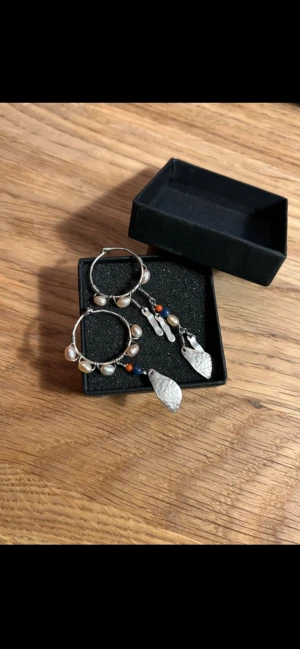 silverörhängen från Maanesten - Säljer ett par handgjorda örhängen i silverfärgad metall med oregelbundna pärlor, blå och orange pärldetaljer samt hamrade silverhängen. Örhängena har en cool bohemisk vibe och är perfekta för dig som gillar unika accessoarer. Ca 5,5 cm