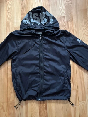Risky vindjacka - Svart vindjacka från Risky med coolt tryck på huvan och ärmen. Jackan har huva med snörning, hel dragkedja framtill och två sidofickor. Materialet är lätt och syntetiskt, perfekt för blåsiga dagar. Passformen är normal och jackan har långa ärmar.