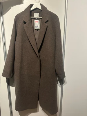 Brun kappa från H&M - Säljer en brun kappa från H&M i oversized rak modell med bred krage och dolda knappar. Jackan är tillverkad i ullblandning och har långa ärmar samt slits bak. Obs: Alla plagg säljs endast fram till 28/12