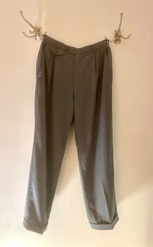 Grå kostymbyxor från Gardeur Homme - Snygga grå kostymbyxor från Gardeur Homme med klassisk rak passform och pressveck. Byxorna har hög midja, knappdetaljer vid fickorna och är tillverkade i ett mjukt bomullsmaterial. Perfekta för en stilren och clean look.