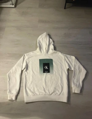 Vit hoodie från Calvin Klein Jeans - Vit hoodie från Calvin Klein Jeans med stor logga i svart och grönt på bröstet. Tröjan har huva, ribbade muddar och är tillverkad i mjuk bomull. Perfekt för chill dagar och har en clean look som funkar till jeans eller joggers.