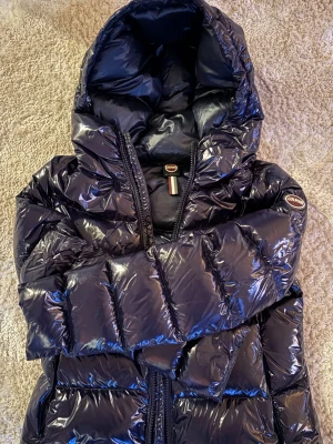 Mörkblå Colmar pufferjacka med huva - Snygg mörkblå pufferjacka från Colmar med glansig finish och stor huva. Jackan har tydliga quiltade sektioner och dragkedja framtill. Colmar-logga på ärmen och insidan. Perfekt för kalla dagar och riktigt stilren look. Passar xs- s och är colmar storleken 44! 