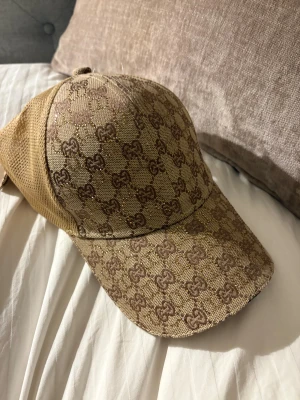 Beige gg keps med glittrigt gg-mönster - Snygg beige keps  med klassiskt gg- mönster i brunt och diskreta glittriga detaljer. Framsidan är i textil och baksidan har mesh för extra ventilation. Perfekt för dig som vill ha en lyxig och trendig accessoar.