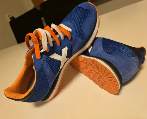 Blå sneakers från Munich strl 43 - Säljer ett par blå sneakers från Munich med vit X-logga på sidan. Skorna har orange och vit snörning, orange innersida och orange sula. Ovandelen är i mesh och mocka för en sportig look. Perfekta för dig som gillar färg och stil.