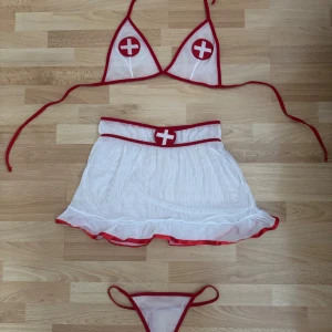 Nya sexiga sjuksköterskan underkläder - Säljer sexiga underkläder i vit mesh med röda kanter och detaljer. Setet består av en behå med snörning, en minikjol med volangkant och ett par stringtrosor. Både toppen och kjolen har röda korsapplikationer framtill för en cool look.  Storleklapp saknas, men den är S/M