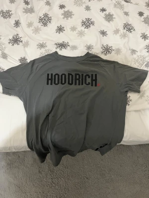 Grå HOODRICH t-shirt med tryck - Säljer en grå t-shirt från HOODRICH med stort svart tryck på bröstet. T-shirten har en klassisk rund hals och korta ärmar, perfekt för en chill streetwear-look. Materialet känns mjukt och skönt mot huden.