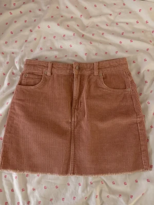 Rosa manchesterkjol från Gina Tricot - Kort kjol i ljusrosa manchester från Gina Tricot. Kjolen har klassisk femficksdesign, bälteshällor och rå kant nedtill. Randigt mönster i tyget och stängs med knapp och dragkedja framtill.