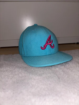 Turkos New Era 59FIFTY keps - Snygg turkos keps från New Era, modell 59FIFTY, med Atlanta Braves A-logga i rosa brodyr framtill och MLB-logga bak. Klassisk formad skärm och stängd baksida. Perfekt för dig som vill sticka ut med färg och sportig vibe. Storlek 7 55,8 cm.