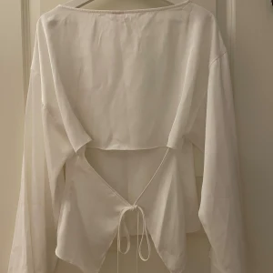 Satinblus med öppen rygg från Nelly - Säljer en vit satin blus från Nelly med långa ärmar och snygg öppen rygg med knytband. Perfekt för dig som gillar stilrena och trendiga plagg. Endast testad