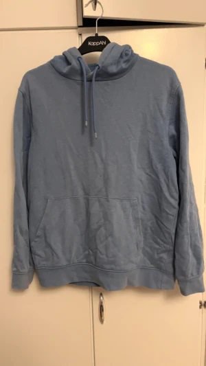 Blå hoodie från Dressmann med huva - Blå hoodie från Dressmann i storlek M. Tröjan har en klassisk huva med snörning och en stor magficka framtill. Tillverkad i mjuk bomull, perfekt för chill dagar. Långärmad och enkel design som funkar till jeans eller joggers.
