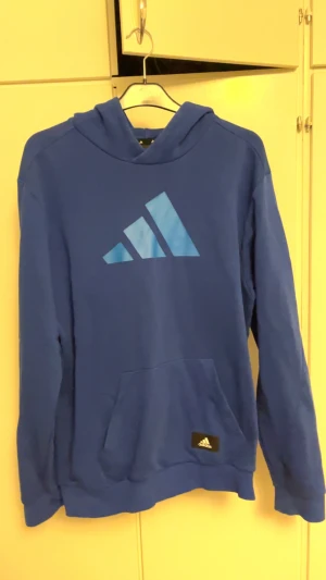Blå hoodie från Adidas med logga - Blå hoodie från Adidas med stor logga i ljusblått på bröstet. Tröjan har huva, magficka och långa ärmar. Tillverkad i mjuk bomullsmix som är skön att ha på sig. Perfekt för chill eller träning, klassisk streetwear-stil.