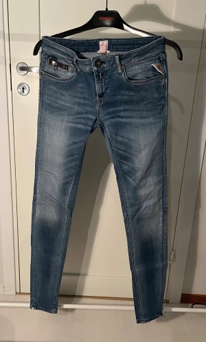 Replay blå jeans slim fit - Snygga blå Replay jeans med slim fit och klassisk femficksdesign. Jeansen har en dragkedjedetalj på ena fickan och subtila slitningar för en cool look. Passformen är smal och materialet är stretchigt denim för extra komfort.