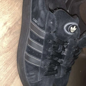 Svarta Adidas Campus sneakers mocka - Säljer ett par svarta Adidas Campus sneakers i mocka med klassiska tre ränder på sidan och Adidas-logga på plösen. Skorna har rund tå, platt sula och snörning med svarta skosnören. Perfekt för dig som gillar stilrena och tidlösa sneakers. Knappt använd